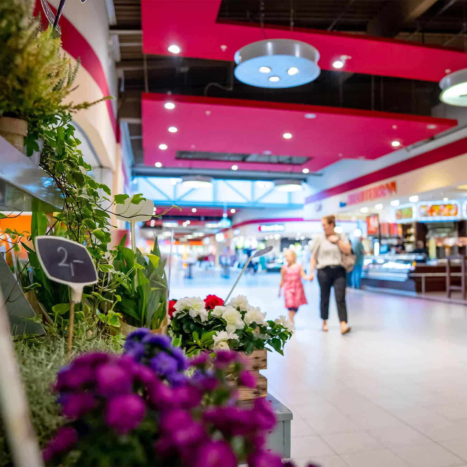 Bunte Blumen in Töpfen stehen links in einem Einkaufszentrum mit mehreren Geschäften im Hintergrund