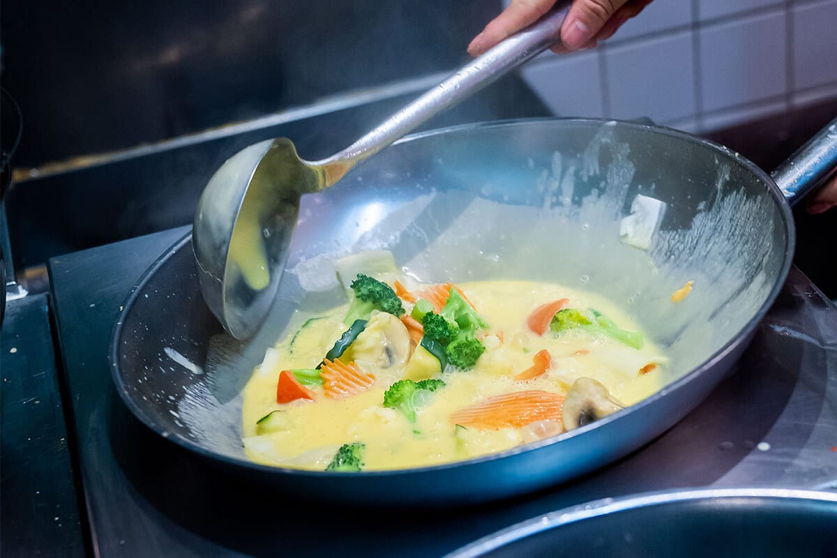 Wok mit cremiger Sauce, die durch eine Hand mit einem Schöpflöffel gerührt wird