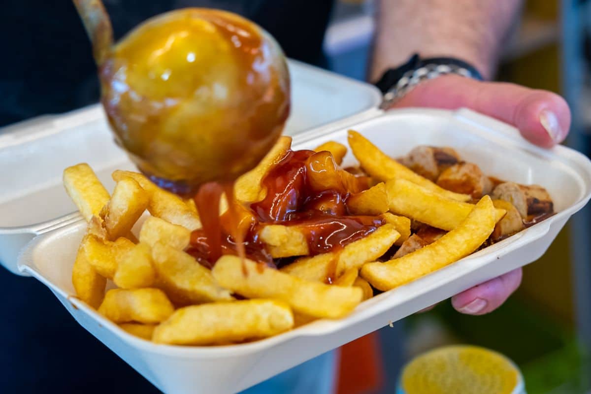 Nahaufnahme einer Schale mit Pommes frites und geschnittener Bratwurst, die mit Currysoße übergossen wird